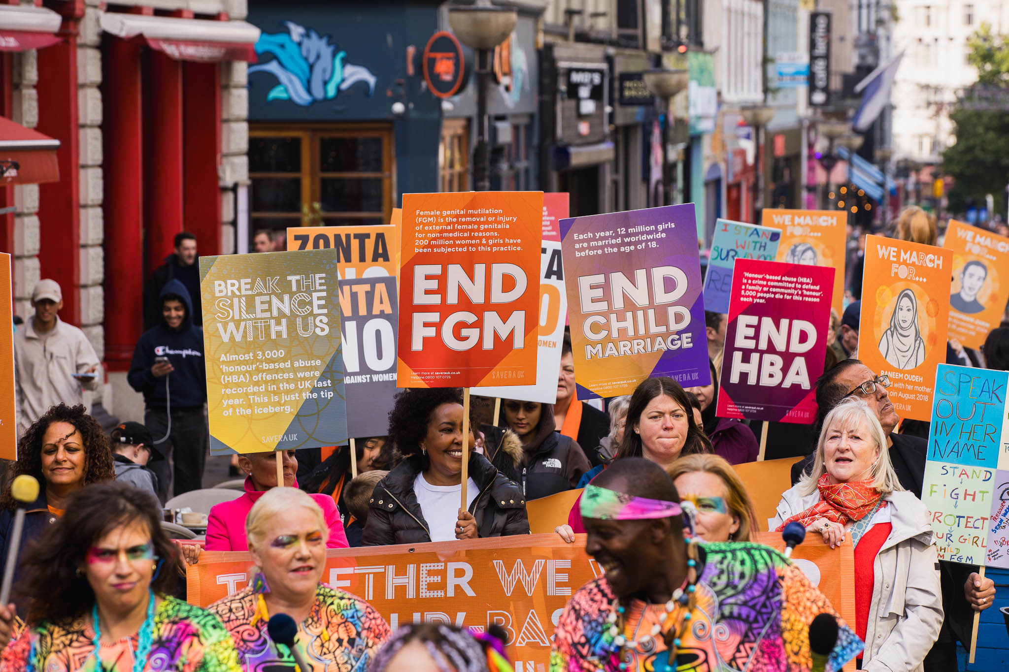 A large crowd marches down a city street holding brightly colored signs calling for an end to honour-based abuse and harmful practices. Posters read messages such as “Break the Silence With Us,” “End FGM,” “End Child Marriage,” and “End HBA.” People of different ages and backgrounds walk together, some wearing vibrant clothing and face paint, showing solidarity and energy as they advocate for human rights.